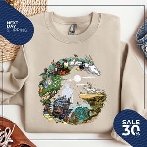 a beige sweatshirt with a colorful illustration of a dragon and various fantastical elements, such as a castle, trees, and people, all set against a whimsical landscape. The sweatshirt is displayed on a white background, and there are other clothing items and accessories visible in the image.