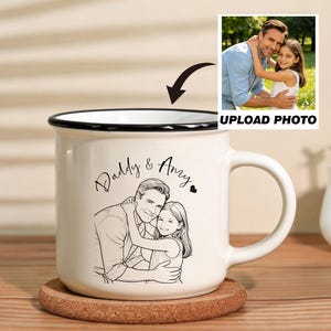 Puede incluir: Una taza de cerámica blanca con borde y asa negros, con un boceto de un padre y un niño abrazándose, con las palabras "Daddy & Amy". Una pequeña foto se muestra encima de la taza con el texto "UPLOAD PHOTO".