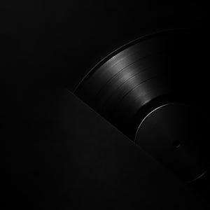 May include: A black vinyl record partially emerging from a black sleeve. The record features concentric circular grooves and a smooth central label area, captured in a low-key lighting style that emphasizes the dark, monochromatic aesthetic.