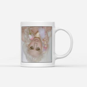 May include: White ceramic coffee mug featuring a portrait of a person with blonde hair, holding a pink flower and a feather. The mug has a standard handle and a glossy finish. The image is centered on the mug.