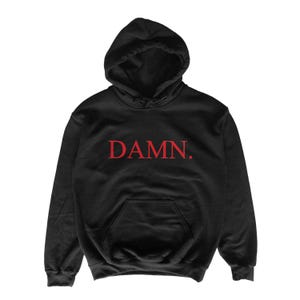 Puede incluir: Sudadera con capucha negra con bolsillo delantero y capucha con cordón. La palabra "DAMN." está impresa en rojo en el pecho. La sudadera está hecha de un material suave.