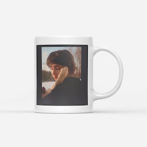 May include: White ceramic mug with a photograph of a person resting their head on their hand. The photograph shows a person with short brown hair, wearing a black top, looking out a window at a snowy landscape.