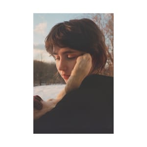 May include: A person with short brown hair, wearing a black top, is gently holding a light-colored object. The background features a snowy landscape and trees under a soft, overcast sky. The image has a warm, muted color palette.