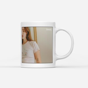 May include: White ceramic mug with a curved handle. The mug features a photograph of a person wearing a white t-shirt with a floral design. The word "Serenity" is printed on the mug.