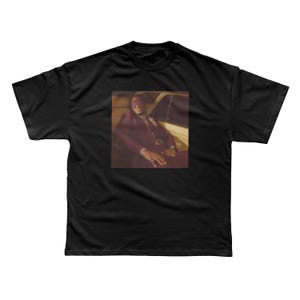 May include: Black t-shirt featuring a framed image of a person in a maroon suit and sunglasses, seated in a car. The image has a warm, sepia-toned color palette. The t-shirt is a classic crew neck style.