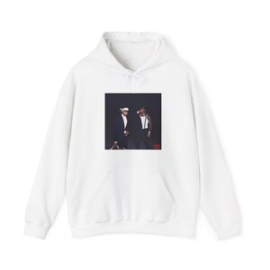 Puede incluir: Sudadera con capucha blanca con bolsillo delantero y un gráfico de dos personas con trajes y gafas de sol. El gráfico es rectangular y está centrado en la parte delantera de la sudadera.