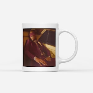 Puede incluir: Taza de cerámica blanca con una fotografía de una persona con un traje burdeos y una venda en los ojos, sentada en un coche. La imagen tiene una estética cálida y vintage. La taza tiene un asa estándar y un acabado brillante.