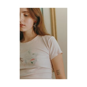 May include: A light pink short-sleeved t-shirt with a faded abstract design. The image also shows a red floral earring and a tattoo on the arm with the letters "A.E.C. G.A.C. A.P.C."