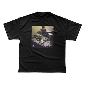 May include: Black t-shirt featuring a graphic print. The print depicts a still life scene with various objects, including a notebook, a pen, and a cup. The text "Lamar" and "80" are visible in the print. The t-shirt has short sleeves and a crew neck.