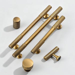 Op de afbeelding: Een verzameling messingkleurige kastbeslagen, waaronder lange stangtrekkers, een cilindrische knop en een T-vormige trekker. De trekkers hebben een geribbeld ontwerp op de handgrepen en zijn gemonteerd op kleine, ronde voetjes. De hardware is op een wit oppervlak geplaatst.