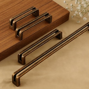 May include: A selection of bronze-coloured cabinet pulls in different lengths. The pulls feature a rectangular design with a central groove, offering a modern, minimalist look. The hardware is presented on a wooden surface, highlighting the warm, metallic finish.