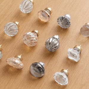 May include: A collection of decorative glass knobs with brass-coloured bases. The knobs display diverse shapes, such as ribbed, faceted, and spherical designs. The glass is clear, frosted, and tinted, providing a variety of textures. They are arranged on a light-coloured wooden surface.