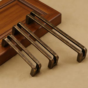 Könnte beinhalten: Vier antike bronzefarbene Schrankgriffe in verschiedenen Größen werden gezeigt. Jeder Griff hat einen rechteckigen Griff mit einem Doppelstangen-Design. Die Griffe sind auf einer hellbraunen Oberfläche angeordnet und zeigen ihre gealterte, Vintage-Ästhetik. Die Hardware ist für Schränke und Schubladen konzipiert.