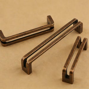 Pode incluir: Três puxadores de armário de cor bronze em tamanhos variados. Os puxadores têm um design retangular com um centro oco e um interior escuro. O hardware é feito de metal e tem uma aparência vintage ou antiga.