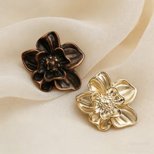 May include: Two decorative flower-shaped metal embellishments. One is bronze-colored, the other is gold. Each flower has detailed petals and a textured center, ideal for craft projects or adding flair to clothing.