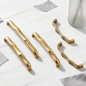 May include: A selection of gold-toned cabinet hardware, comprising four bamboo-style handles and two decorative pulls. The hardware is displayed on a white wooden surface, with soft lighting. The overall style is modern and elegant, suitable for various interior designs.