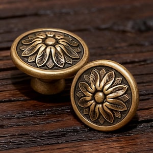 May include: Two antique brass cabinet knobs with a floral design. The knobs are round with a raised flower pattern and a dark, aged finish. They are set against a dark wood background.