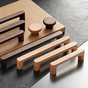 May include: A selection of wooden cabinet handles and knobs in different sizes and finishes. The pulls are dark brown and natural wood, displayed on a light wooden board. The background is dark.