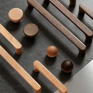 Może przedstawiać: Asortyment drewnianych okuć do szafek, w tym gałki i uchwyty. Gałki są okrągłe, a uchwyty prostokątne z zaokrąglonymi krawędziami. Odcienie drewna wahają się od jasnobrązowego do ciemnobrązowego, ułożone na ciemnoszarej powierzchni.