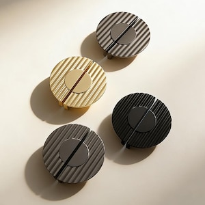 May include: Four contemporary cabinet pulls in gold, black, and grey. Each pull has a circular design with a central disc and a grooved, ridged outer ring. The pulls are arranged on a neutral background, highlighting their modern aesthetic.