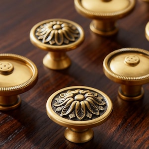 May include: Close-up of several antique brass cabinet knobs. Some knobs feature a floral design, while others have a simple, round shape with a beaded edge. The knobs are set against a dark wood surface.