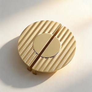 Puede incluir: Un tirador de armario circular de color dorado con un anillo exterior acanalado y un centro circular dividido. El herraje tiene un diseño moderno y está hecho de metal. El tirador se muestra sobre un fondo blanco.
