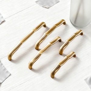 May include: Six gold-colored cabinet pulls with a bamboo-inspired design are arranged on a white wooden surface. The pulls have a curved handle and a textured, segmented body. A frosted glass vase is visible in the background.