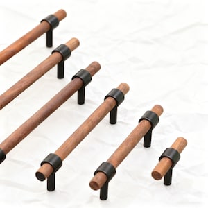 Może przedstawiać: Zestaw drewnianych uchwytów do szafek. Każdy uchwyt ma cylindryczny drewniany uchwyt, czarny metalowy pierścień i czarną metalową podstawę. Drewno ma ciepły brązowy odcień, a metal jest matowy czarny.