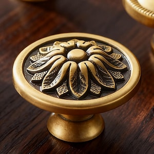 Op de afbeelding: Decoratieve messing kastknop met een bloemmotief. De knop heeft een gedetailleerde bloem in het midden, omgeven door een cirkelvormig frame. De afwerking is een warme, antieke goudkleur, die een vleugje vintage charme toevoegt.