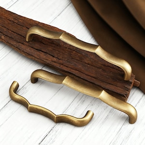 Może przedstawiać: Trzy uchwyty do szafek z antycznego mosiądzu o zakrzywionym kształcie. Uchwyty mają ciepły, złoty kolor i lekko postarzaną powierzchnię. Ułożone są na kawałku zwietrzałego drewna.