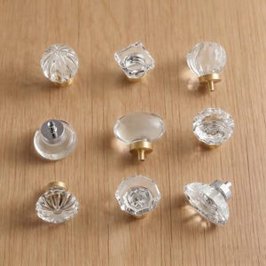 Puede incluir: Nueve pomos de armario de vidrio transparente con varios diseños, incluyendo formas acanaladas, facetadas y ovaladas. Cada pomo tiene una base dorada y un tornillo plateado. Los pomos están dispuestos sobre una superficie de madera marrón claro.