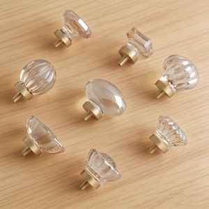 May include: A collection of clear glass cabinet knobs with brass-colored bases. The knobs have various shapes, including round, oval, and faceted designs. The knobs are arranged on a light-colored wooden surface.