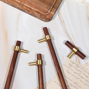 May include: Four dark brown wooden cabinet pulls with gold-coloured brass accents. The pulls are cylindrical in shape. A wooden chopping board is visible in the background. The image is set on a marble surface.