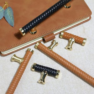 May include: A collection of decorative cabinet pulls with braided leather handles in black and various shades of brown. The pulls have gold-toned metal accents and are arranged near a brown leather notebook.