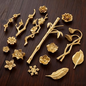 May include: A collection of gold-colored cabinet hardware, including floral and botanical designs. The hardware features various shapes, such as flowers, branches, bamboo, and leaves, all with intricate detailing. The pieces are arranged on a dark wooden surface.
