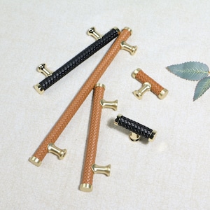 May include: A collection of cabinet pulls with woven leather handles in black and various shades of brown. The pulls have gold-colored, T-shaped mounting hardware. The cabinet pulls are designed for furniture and home decor.