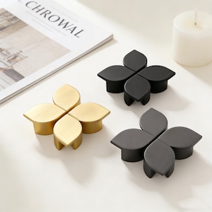 May include: Three decorative cabinet knobs in a flower shape. One is gold, one is black, and one is dark gray. The knobs are on a white surface with a magazine and a candle in the background.