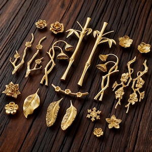May include: A collection of gold-colored decorative hardware, including floral, bamboo, and leaf-shaped drawer pulls and knobs. The hardware is arranged on a dark wood surface, showcasing various designs and sizes for home decor.