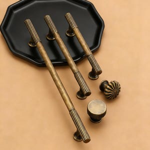 Può includere: Un set di ferramenta per mobili in ottone antico, tra cui tre lunghe maniglie a barra e due stili di pomolo. Le maniglie e i pomoli hanno un design a coste e sono esposti su un vassoio nero su sfondo neutro.
