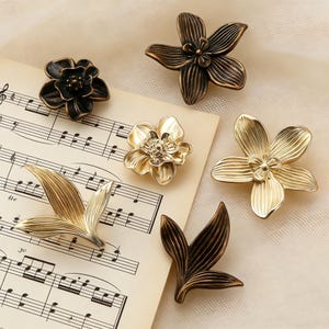 Könnte beinhalten: Eine Sammlung dekorativer floraler Schrankknöpfe und -griffe in verschiedenen metallischen Ausführungen. Die Knöpfe umfassen Blumen- und Blattdesigns in Gold, Bronze und Schwarz. Die Knöpfe sind auf einem Notenblatt angeordnet.