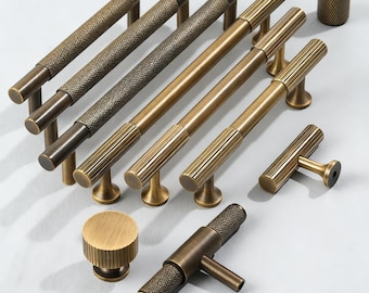 May include: A collection of bronze-toned cabinet hardware, including various handle styles and a knob. The handles feature textured and grooved designs, with a mix of bar and T-shaped pulls. The hardware is arranged on a light surface.