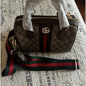 Puede incluir: Un bolso marr&oacute;n con un patr&oacute;n repetitivo y una franja roja y verde en el centro. El bolso tiene una cremallera dorada y una correa desmontable con rayas rojas y verdes. El bolso tiene un logotipo "G" dorado.