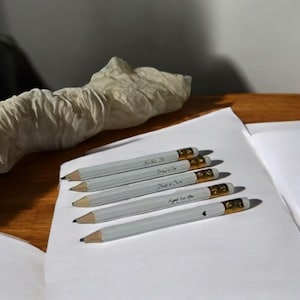 May include: Five gray pencils with gold-colored metal bands and white erasers are arranged on a white sheet of paper. Each pencil has a different inscription in black. The pencils are sharpened and ready for use.