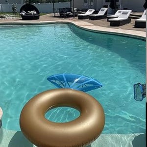 May include: A gold-colored inflatable pool tube floats in a clear blue swimming pool. A blue diamond-shaped pool float is also in the water. The pool has a concrete border with lounge chairs and a seating area in the background.