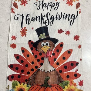 Puede incluir: Bandera decorativa con temática de Acción de Gracias con un pavo de dibujos animados con sombrero de copa, una calabaza y detalles florales. La bandera dice "Happy Thanksgiving" en escritura negra, rodeada de hojas de otoño.