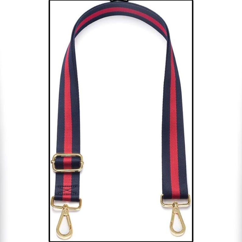Puede incluir: Una correa de bolso a rayas azul marino y rojo con herrajes met&aacute;licos dorados. La correa presenta una franja roja central flanqueada por rayas azul marino. Se ven cierres y una hebilla de metal dorado.