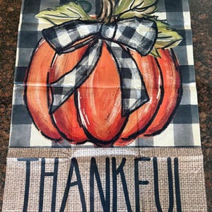 Könnte beinhalten: Ein dekoratives Küchentuch zum Thema Herbst. Es zeigt einen gemalten orangefarbenen Kürbis mit einer schwarz-weiß karierten Schleife und einem Banner im Leinenstil mit der Aufschrift "THANKFUL" in schwarzen Buchstaben. Der Hintergrund ist ein schwarz-weiß kariertes Muster.