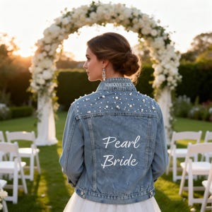 Puede incluir: Una chaqueta de mezclilla azul claro con adornos de perlas blancas en la parte superior de la espalda y los hombros. El cuello muestra el texto en números romanos MMXXVI.V.XX y la espalda tiene el texto bordado en blanco Pearl Bride.