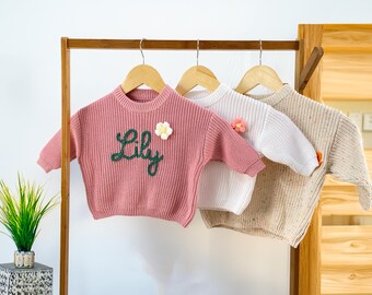 May include: Three children's sweaters in pink, white, and beige hang on a wooden rack. The pink sweater has the name "Lily" embroidered in green and a flower applique. The white and beige sweaters also have flower appliques.