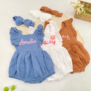May include: Three sleeveless cotton rompers in blue, white, and rust, each featuring ruffled shoulder straps and personalized embroidery. The blue romper says Amelia, the white one says Daisy with a flower, and the rust one says Lucy. Includes matching headbands.